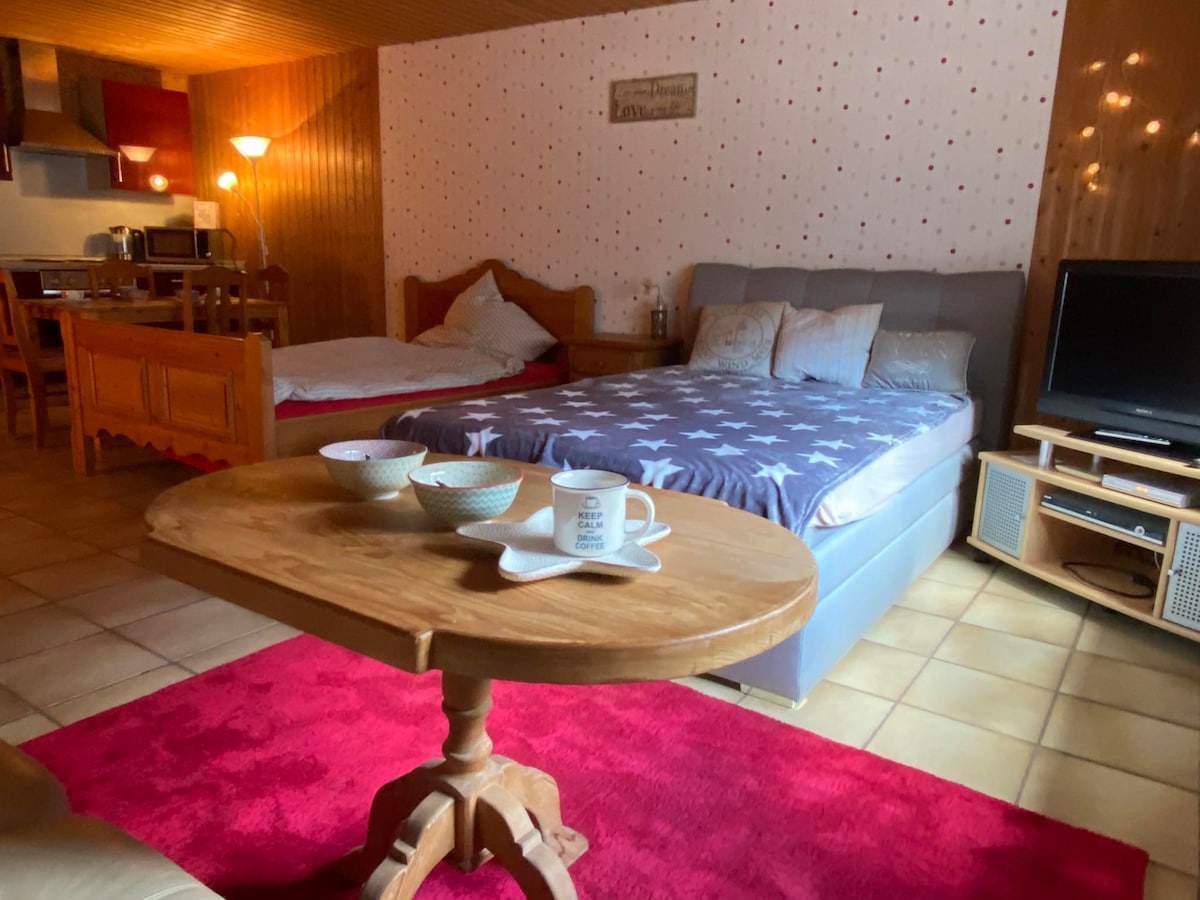 A spacious room features two double beds with a decorative bedspread, complemented by a cozy seating area. A wooden table is set with bowls and mugs. A flat-screen TV sits on a media unit, with soft lighting enhancing the warmth of the space.