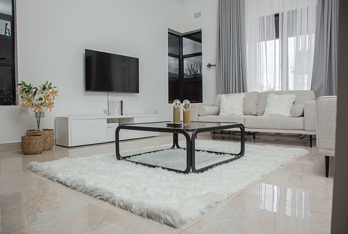 A spacious living area is presented with a light-colored sofa adorned with soft pillows. A modern coffee table sits atop a plush rug. A large television is mounted on the wall, and natural light floods the room through expansive windows.