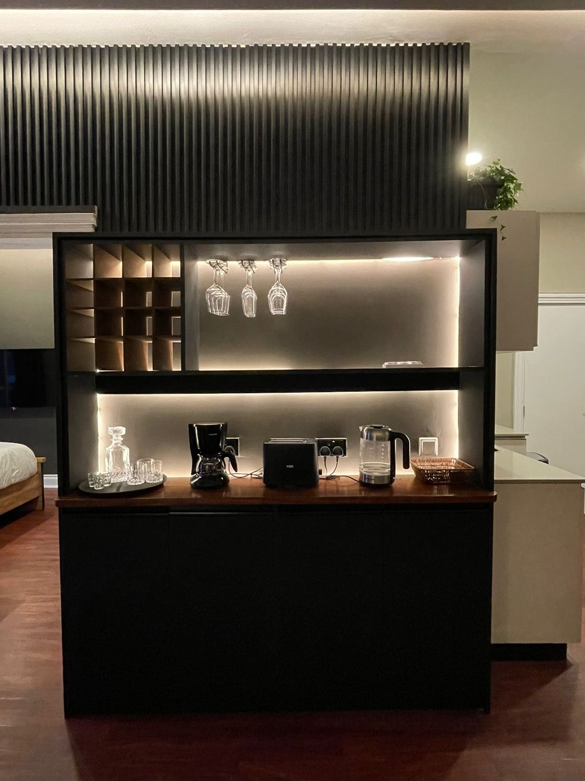 A sleek coffee station is displayed against a dark accent wall, featuring illuminated shelving that showcases glasses and coffee-making equipment. The counter includes a coffee maker, a water purifier, and accessories for a convenient beverage-making experience.