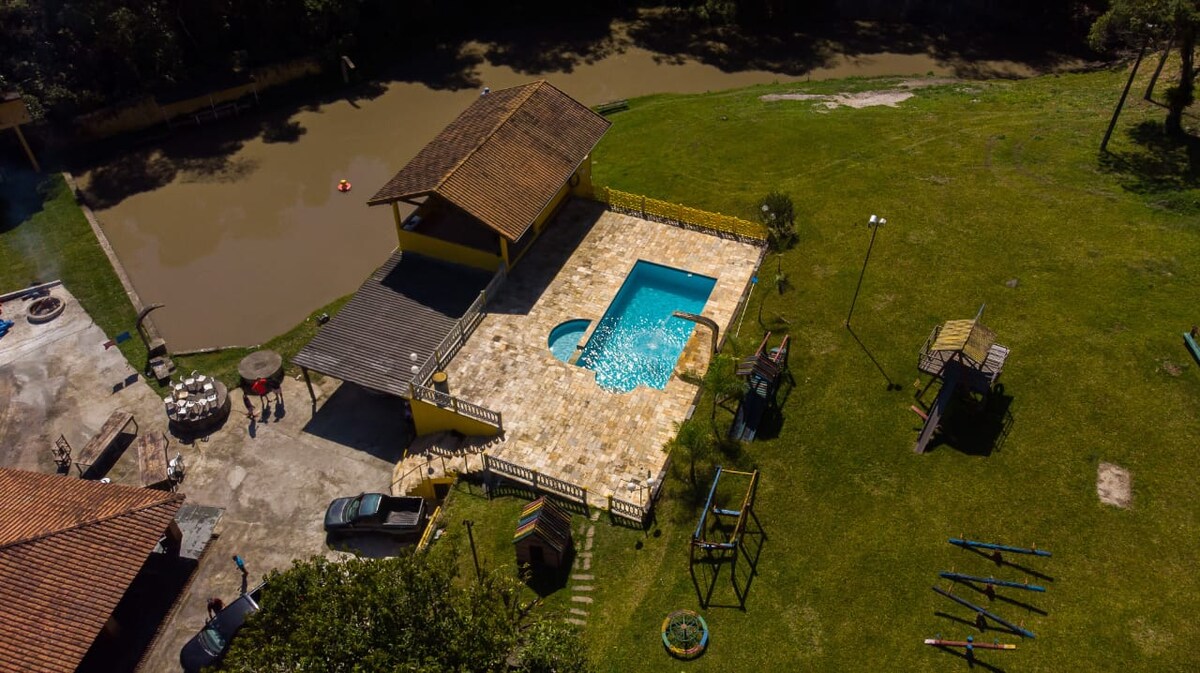An aerial view captures a spacious outdoor area featuring a clear swimming pool surrounded by a tiled deck. Nearby, a playground with various structures is visible on a green lawn, alongside several kayaks positioned near the water's edge.