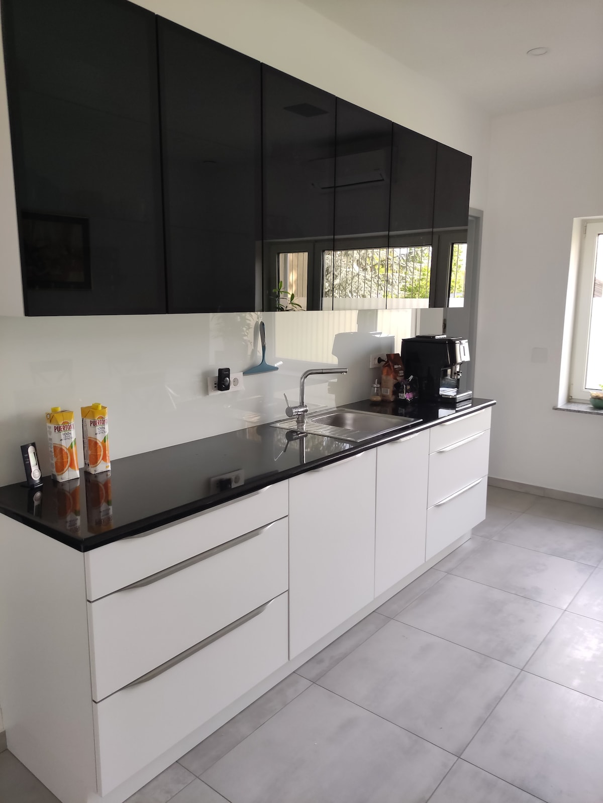 A modern kitchen features sleek black cabinetry and a glossy white countertop. A stainless steel sink is positioned beneath a window, allowing natural light to enter. A coffee maker and various kitchen accessories are visible, contributing to a functional cooking space.