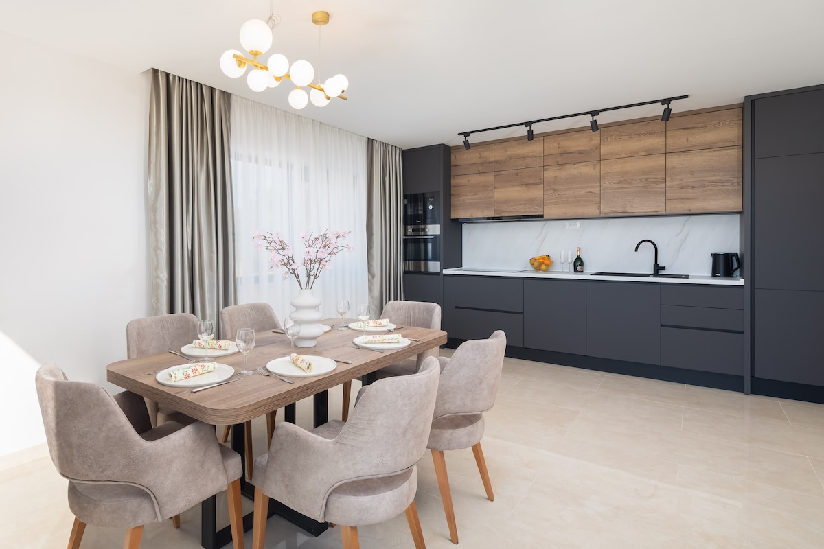 A modern dining area features a wooden table set for six, surrounded by upholstered chairs. Soft natural light filters through sheer curtains, illuminating the sleek cabinetry in the background. An elegant light fixture adds a touch of style to the room.
