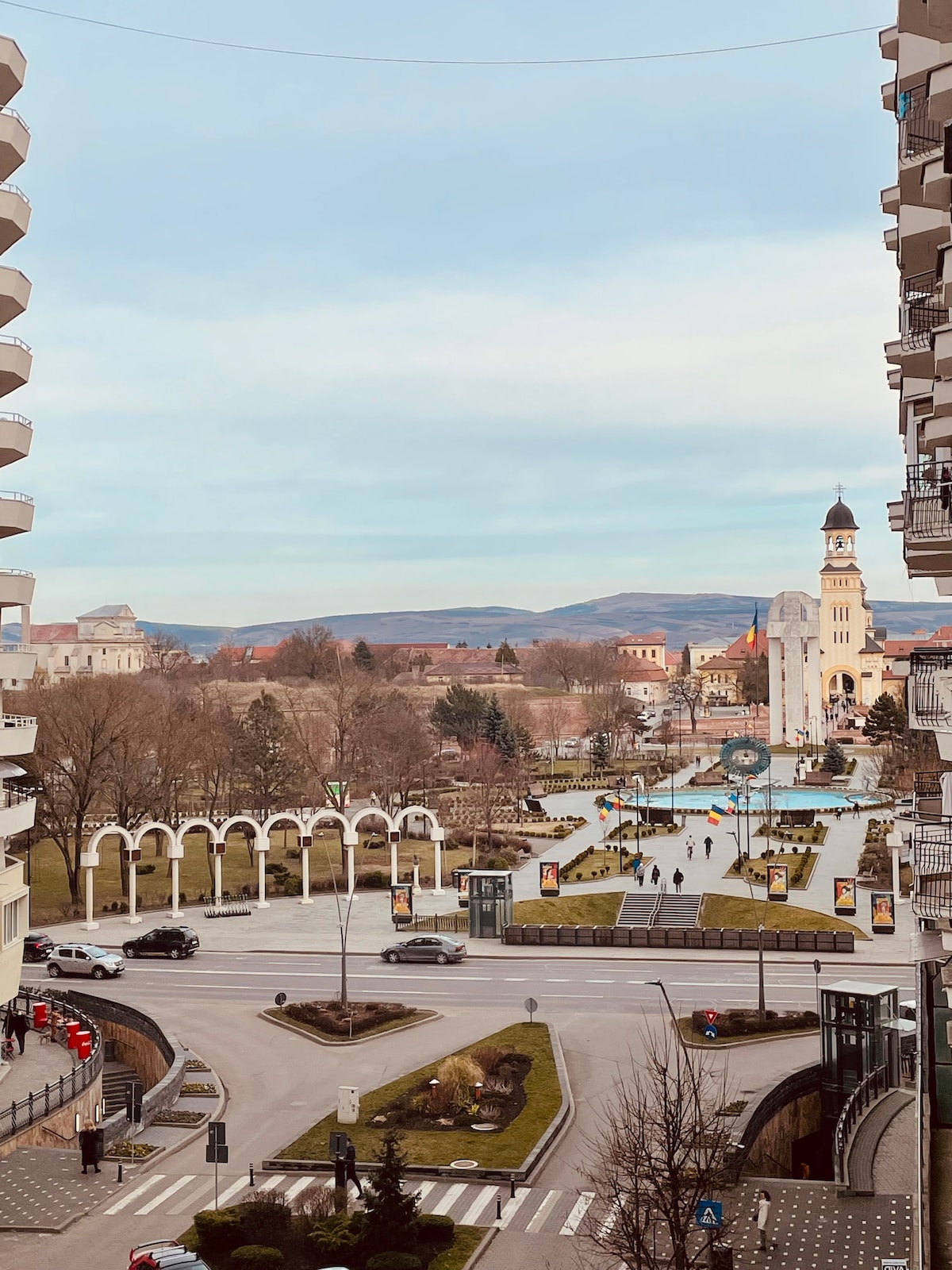 A view from the balcony showcases a lively plaza with people walking, framed by historic architecture. The clock tower is prominent in the background, along with a scenic backdrop of rolling hills under a soft blue sky.