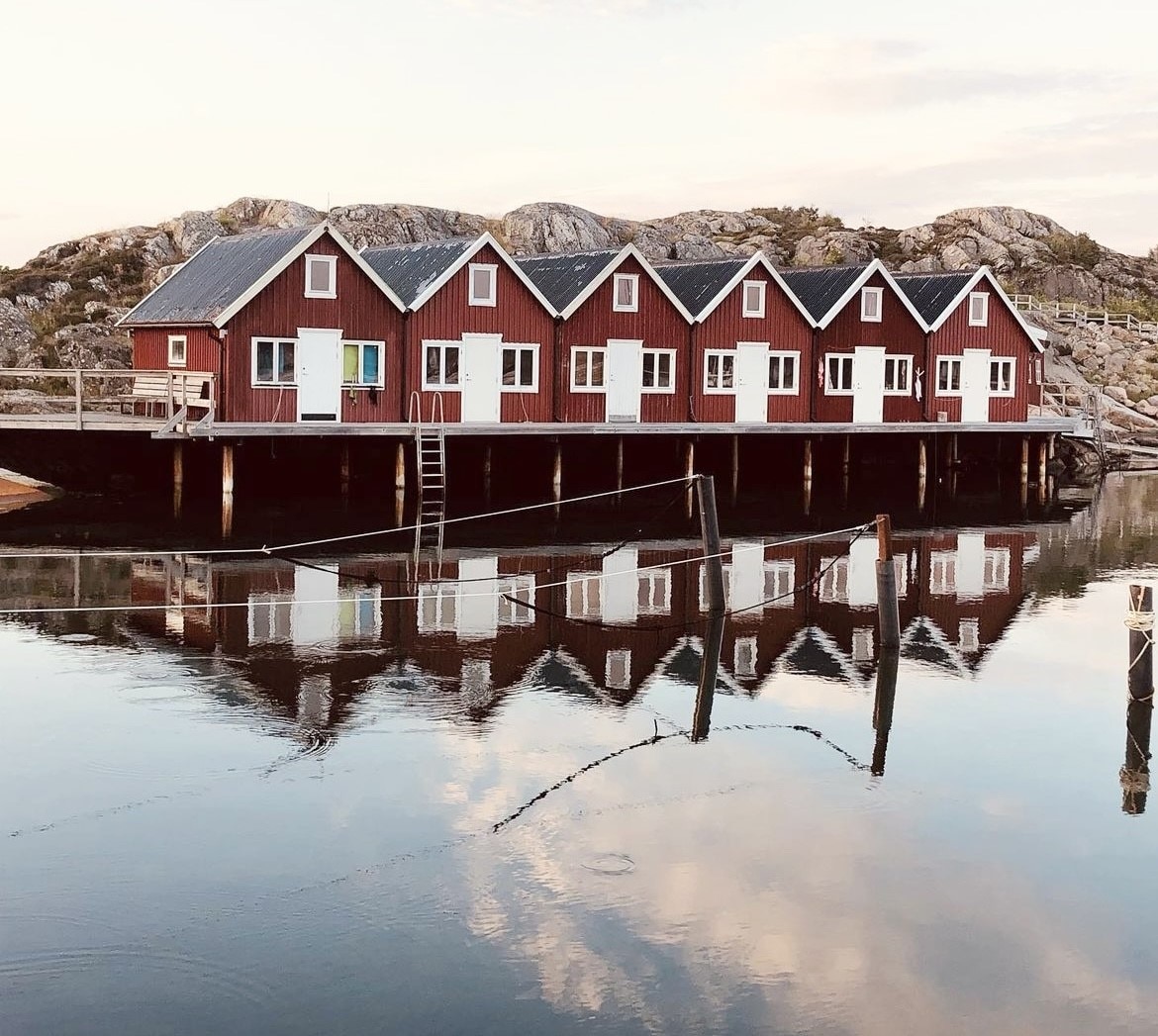 A series of rustic red buildings with white doors are reflected in calm waters, creating a symmetrical appearance. The surrounding rocky landscape and soft clouds in the sky add to the serene setting, suggesting a peaceful retreat by the water.