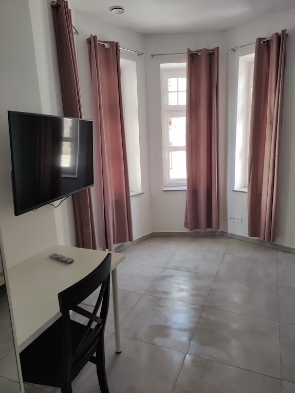 A bright corner of a room is shown, featuring light grey tiled flooring and large windows dressed with soft pink curtains. A small desk with a black chair is positioned beside the wall-mounted TV, providing a functional workspace.