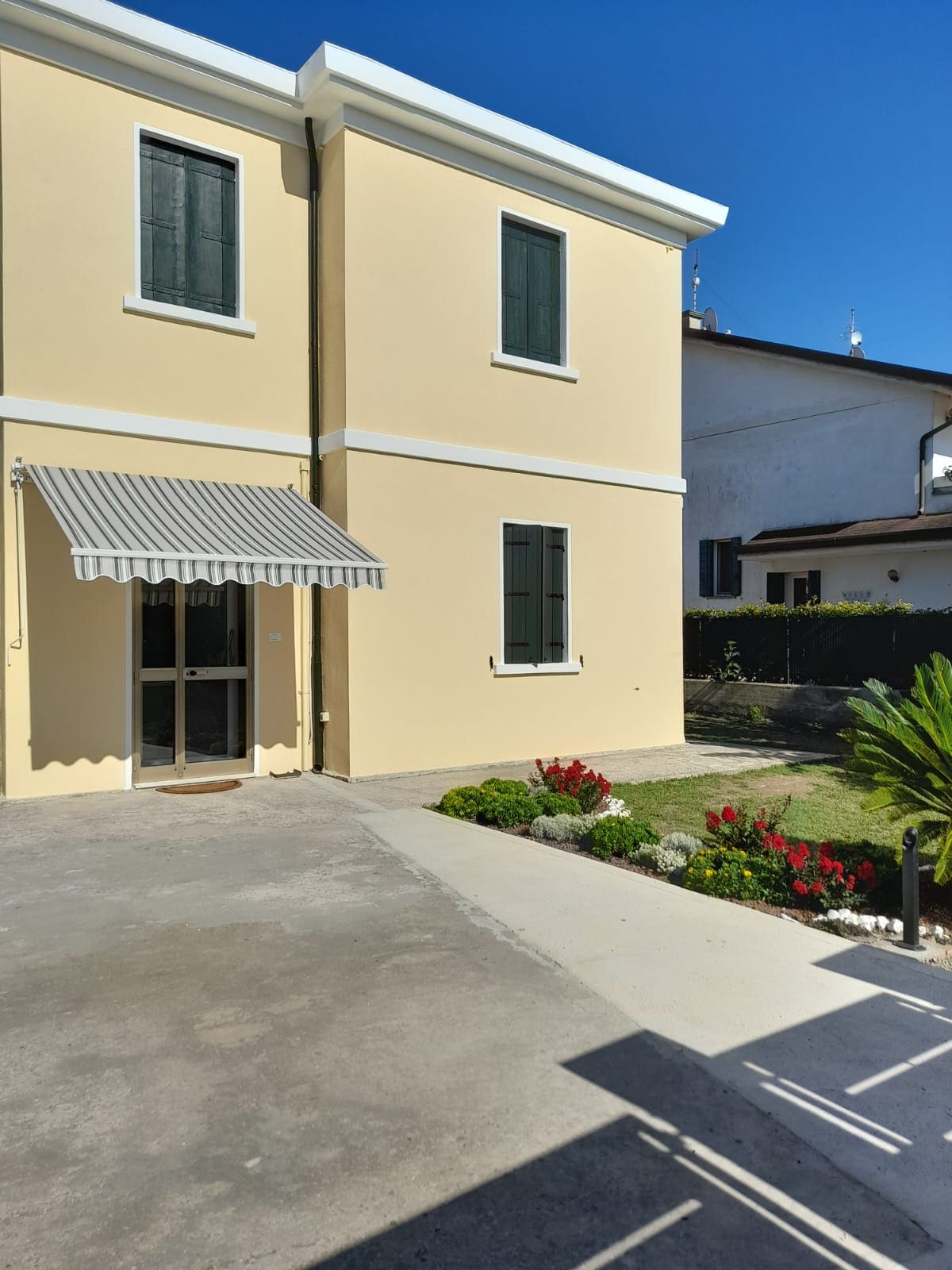 A two-story building features a light-colored exterior with green shutters. A striped awning extends over the entrance, and a small garden with colorful flowers and neatly trimmed shrubs is visible on the side. A paved pathway leads to the entrance.