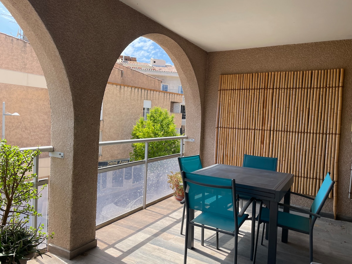 A balcony area is presented with an outdoor dining table surrounded by turquoise chairs. Arched openings provide a sense of space and natural light. A bamboo wall feature adds texture, while potted greenery enhances the inviting character of this outdoor setting.