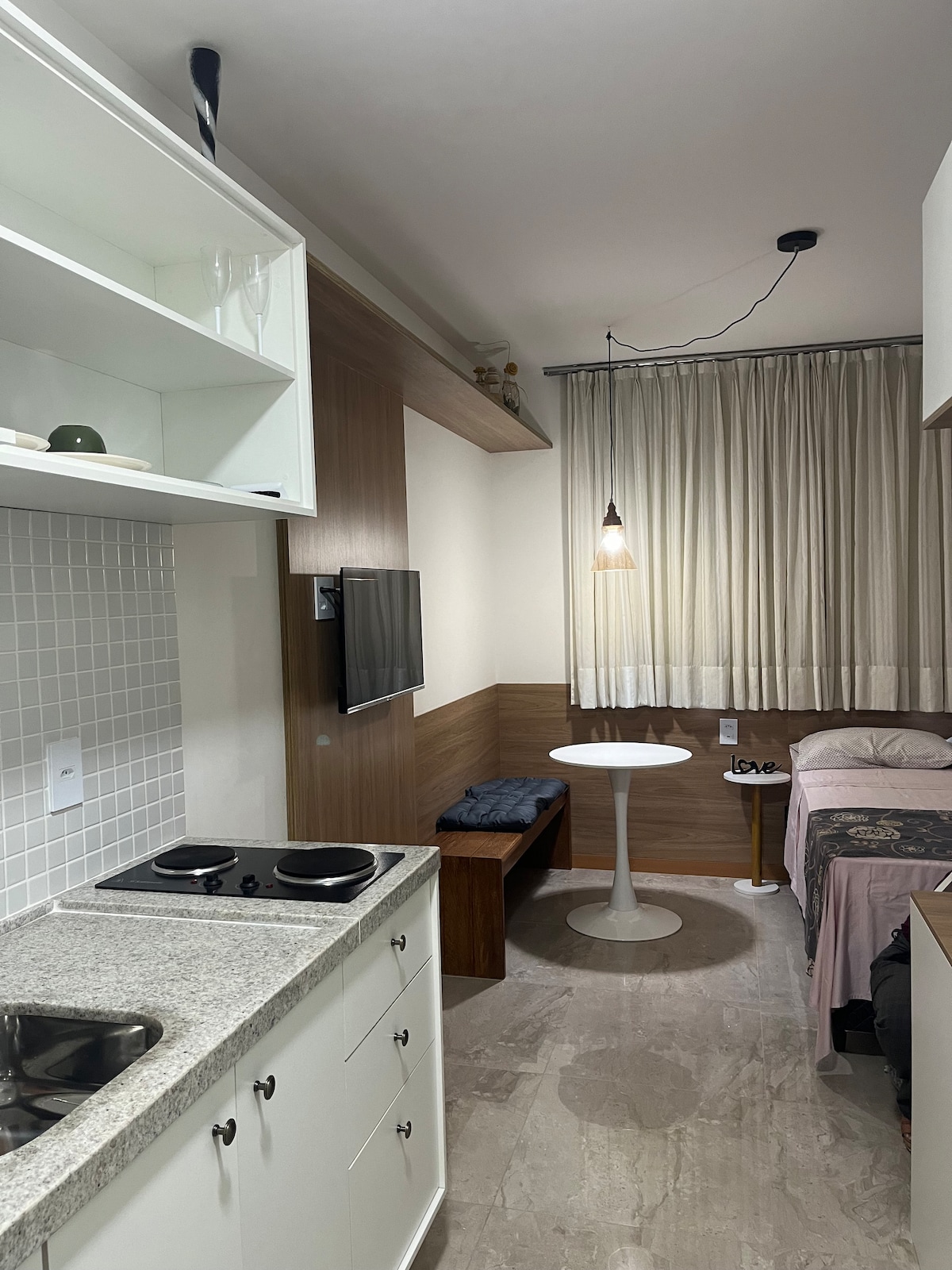 A compact studio space features a small kitchenette with a granite countertop and two burner stovetop. A round dining table is positioned nearby, while a cozy sleeping area offers a bed with soft linens. Natural light enters through sheer curtains, adding warmth to the environment.