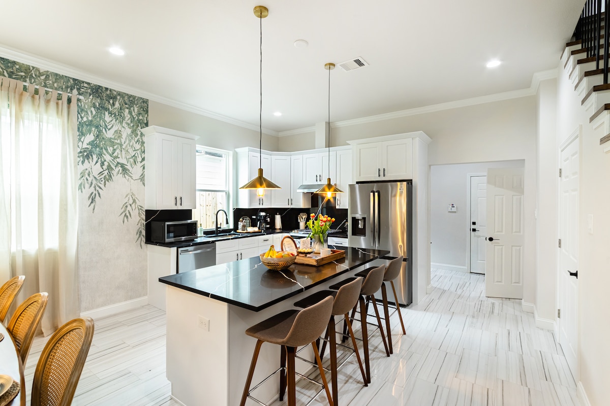 A spacious kitchen features an island with seating for four, complemented by pendant lighting above. Modern appliances are neatly arranged alongside white cabinetry. A dining area is visible in the foreground, with greenery wallpaper enhancing the natural ambiance. Large windows allow ample natural light.