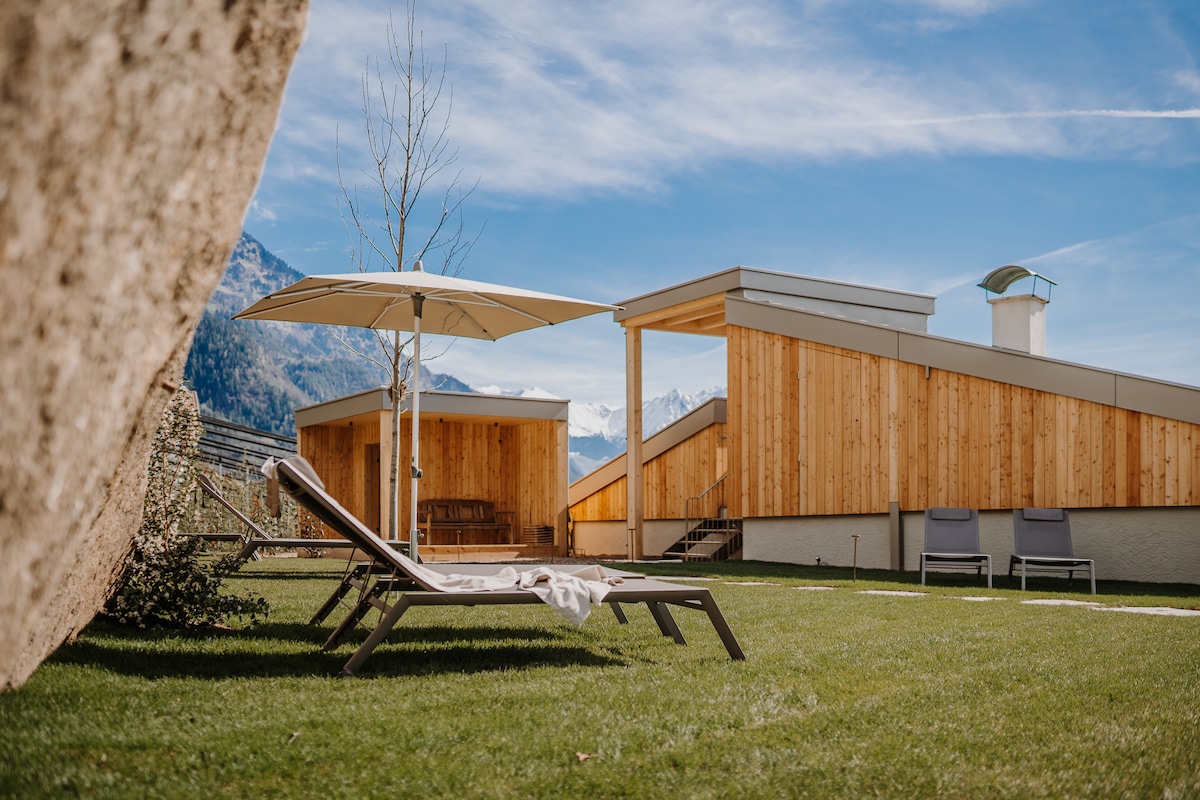A tranquil outdoor space features a sun lounger beneath a large umbrella on a manicured lawn. The wooden structures in the background exhibit a contemporary design, framed by surrounding mountains and a clear blue sky. The scene suggests a relaxing retreat.