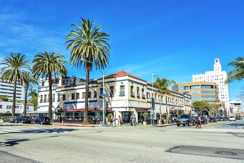 A vibrant street scene is visible with palm trees lining the sidewalks. A multi-story building with a red-tiled roof features outdoor seating below large awnings. Cars are parked along the street, and clear blue skies fill the backdrop.