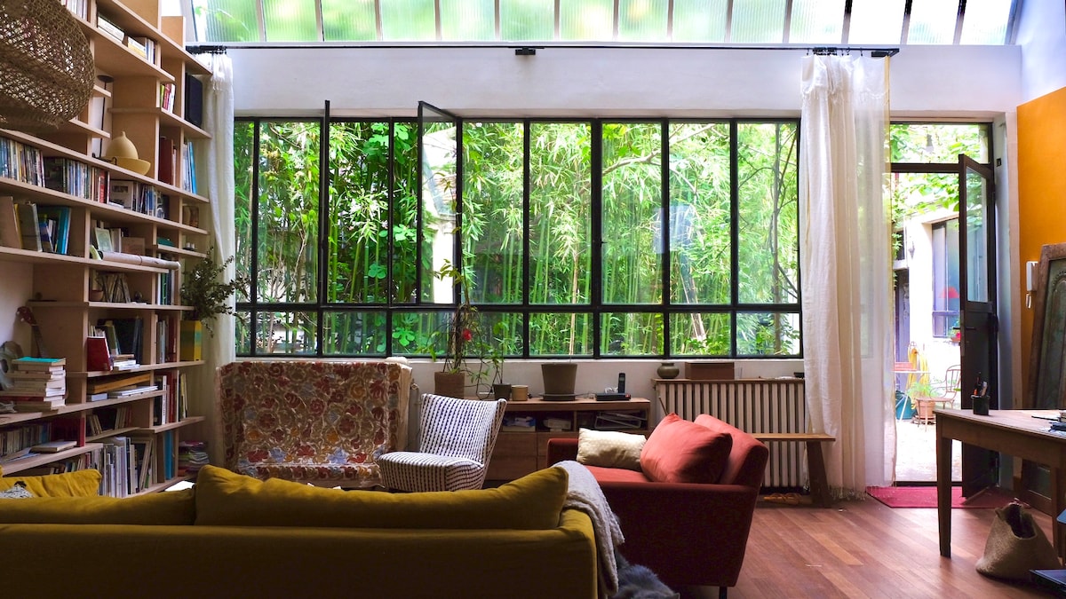 A spacious living area is illuminated by natural light streaming through large glass windows. Various plants, including bamboo, are visible outside, enhancing the green view. Comfortable seating options include a colorful sofa and chairs, while a bookshelf and a coffee table add functional touches.