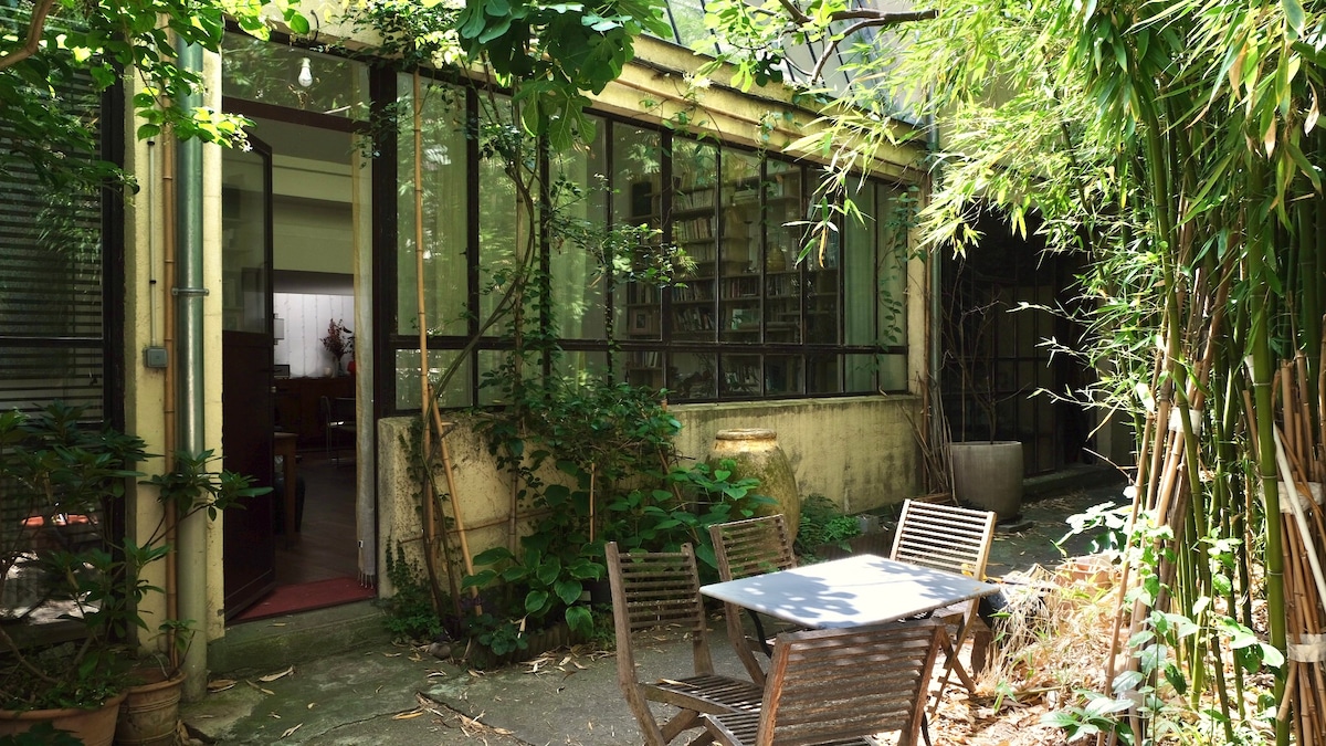 A charming outdoor space features a small wooden table with two chairs, surrounded by abundant greenery including bamboo and other plants. Large windows reveal glimpses of the interior, enhancing the connection between indoor and outdoor areas.