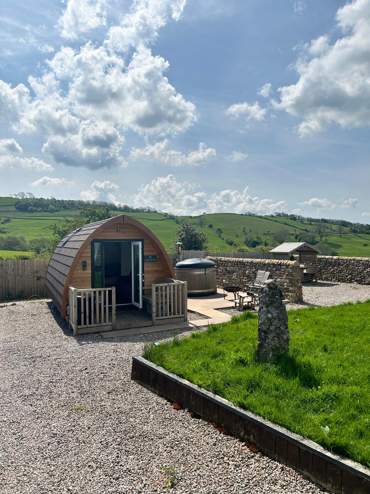 A glamping pod nestled in the Eden Valley countryside is framed by lush green hills. The pod features a wooden deck with railing and is accompanied by a private outdoor hot tub. A gravel pathway leads to a stone feature and well-maintained grassy areas.