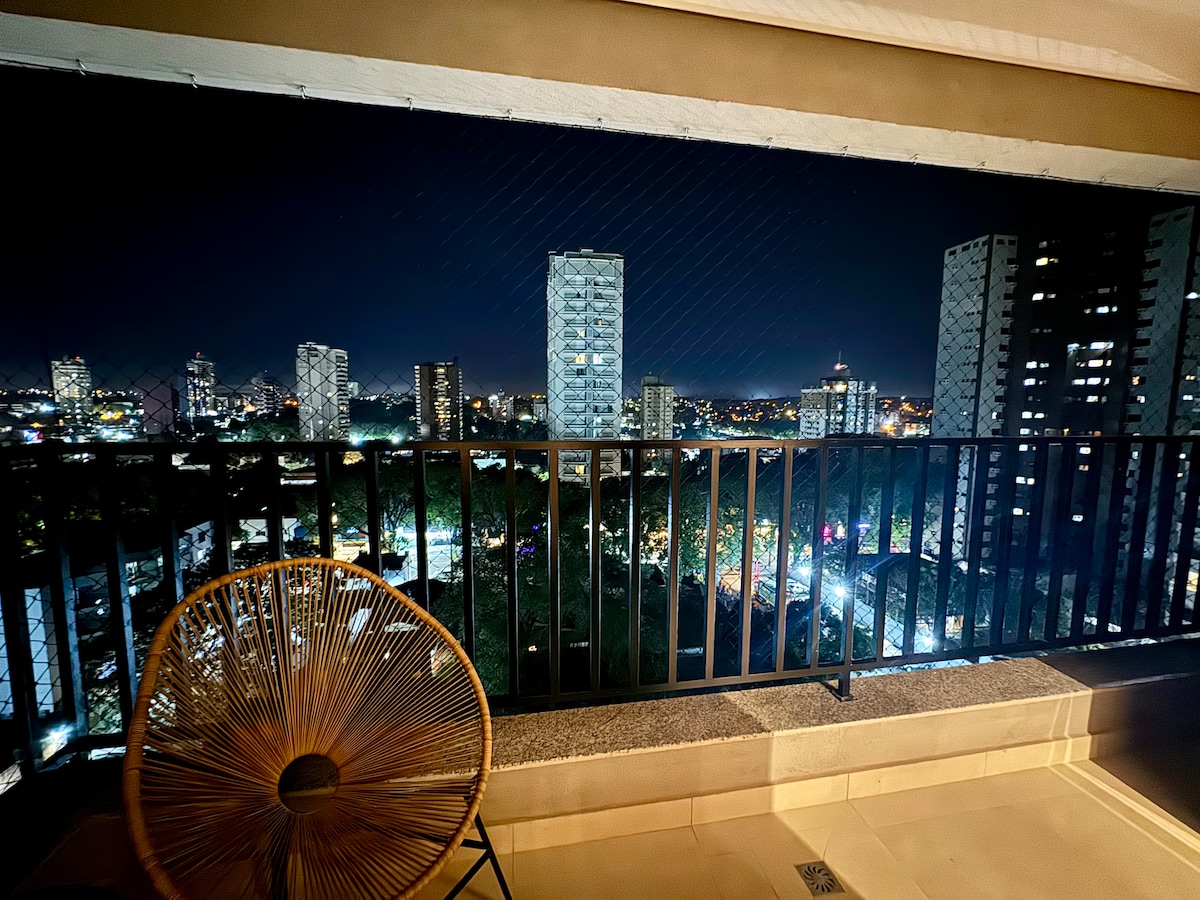 A balcony area is depicted at night, featuring a modern railing and a round chair. The city skyline is visible, with illuminated buildings and a blend of lights creating a serene atmosphere. The expansive view showcases the vibrancy of the surrounding urban landscape.