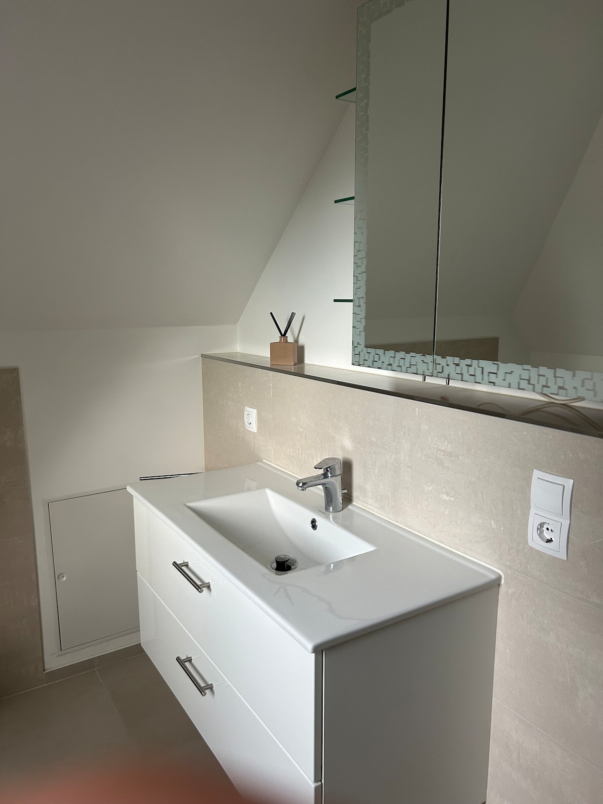 A modern bathroom features a sleek white vanity with two storage drawers and a rectangular basin. Above, a frameless mirror reflects natural light, while decorative items sit on a shelf nearby. A subtle color palette creates a calm, clean atmosphere.