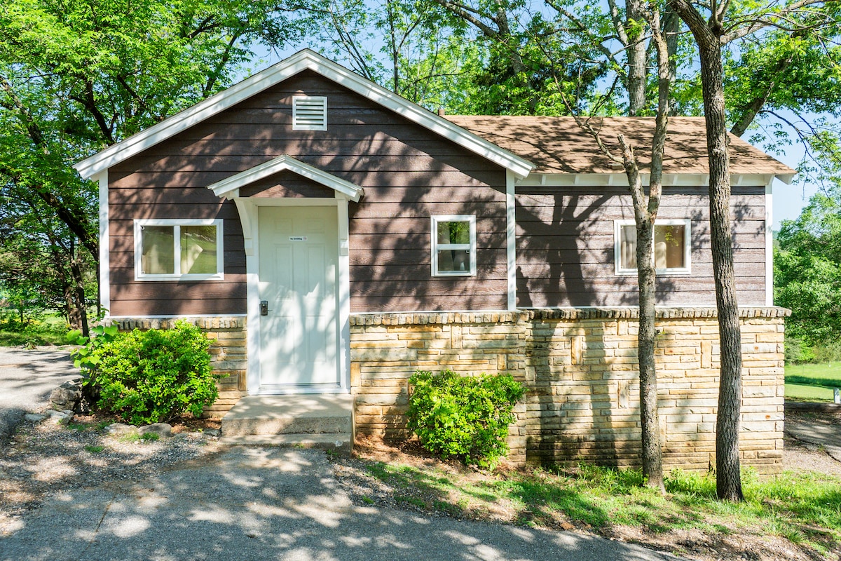 The exterior of a quaint cabin is displayed, featuring dark wood siding contrasted with a stone foundation. A white door with a small window is centered, flanked by additional square windows. Green shrubs line the walkway, and sunlight filters through trees surrounding the cabin.
