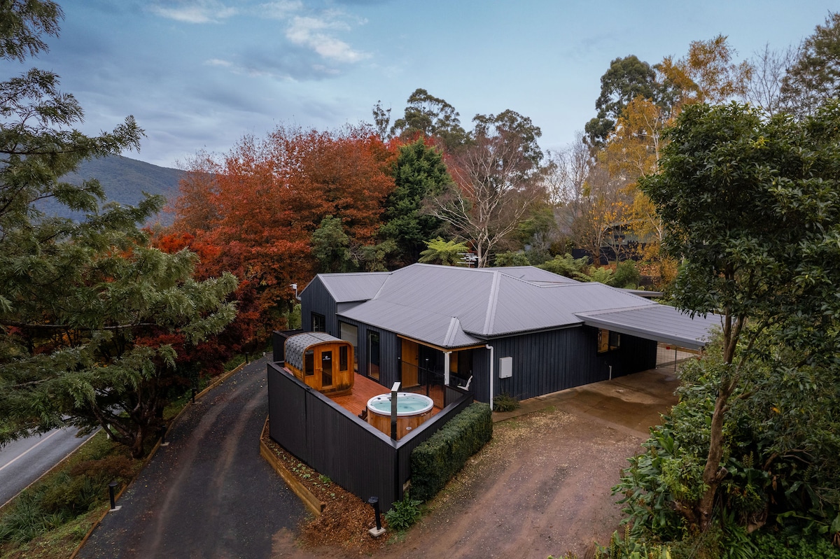 An inviting contemporary retreat is nestled among vibrant autumn foliage, showcasing a private outdoor spa on a wooden deck. The structure is modern, featuring a spacious layout and surrounded by lush greenery, blending seamlessly with the tranquil natural environment.