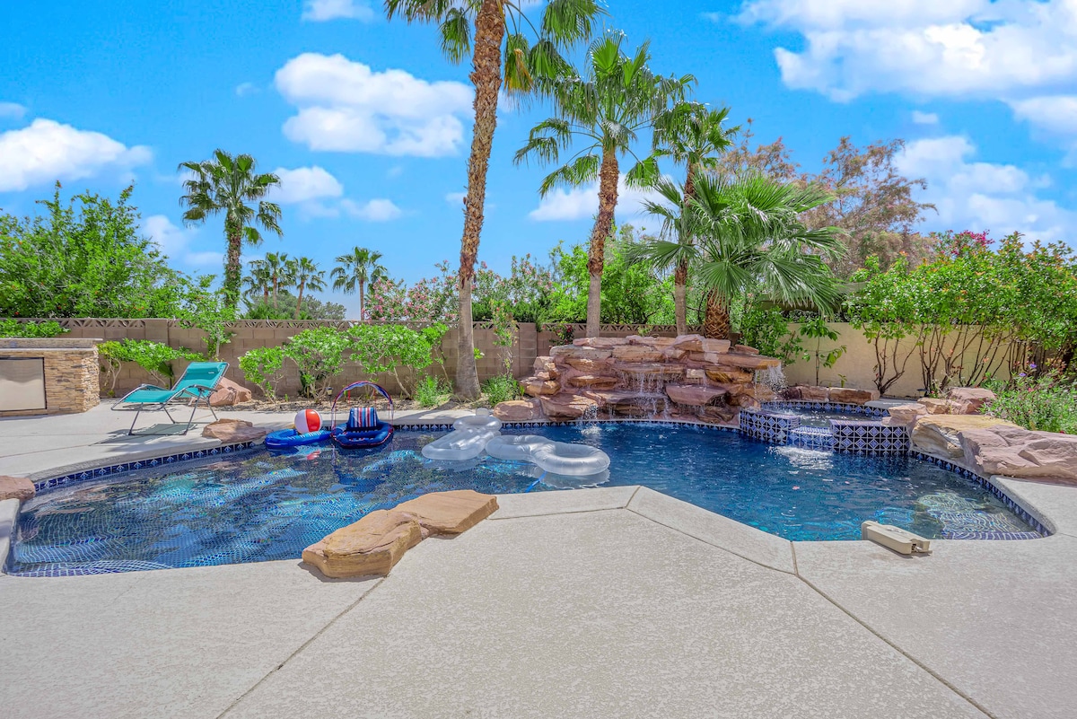 A private heated pool is surrounded by lush greenery and palm trees, with a small waterfall feature cascading into the water. Comfortable lounge chairs and floating accessories are placed on the pool deck, providing a serene space for relaxation under a bright blue sky.