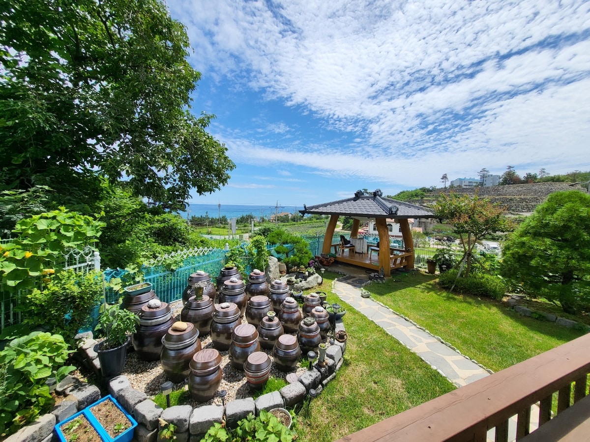 A garden is visible, featuring traditional pottery arranged in a circular formation. A wooden pavilion is positioned nearby, offering a shaded seating area. Lush greenery surrounds the space, with expansive ocean views in the background and a partially cloudy sky providing natural light.