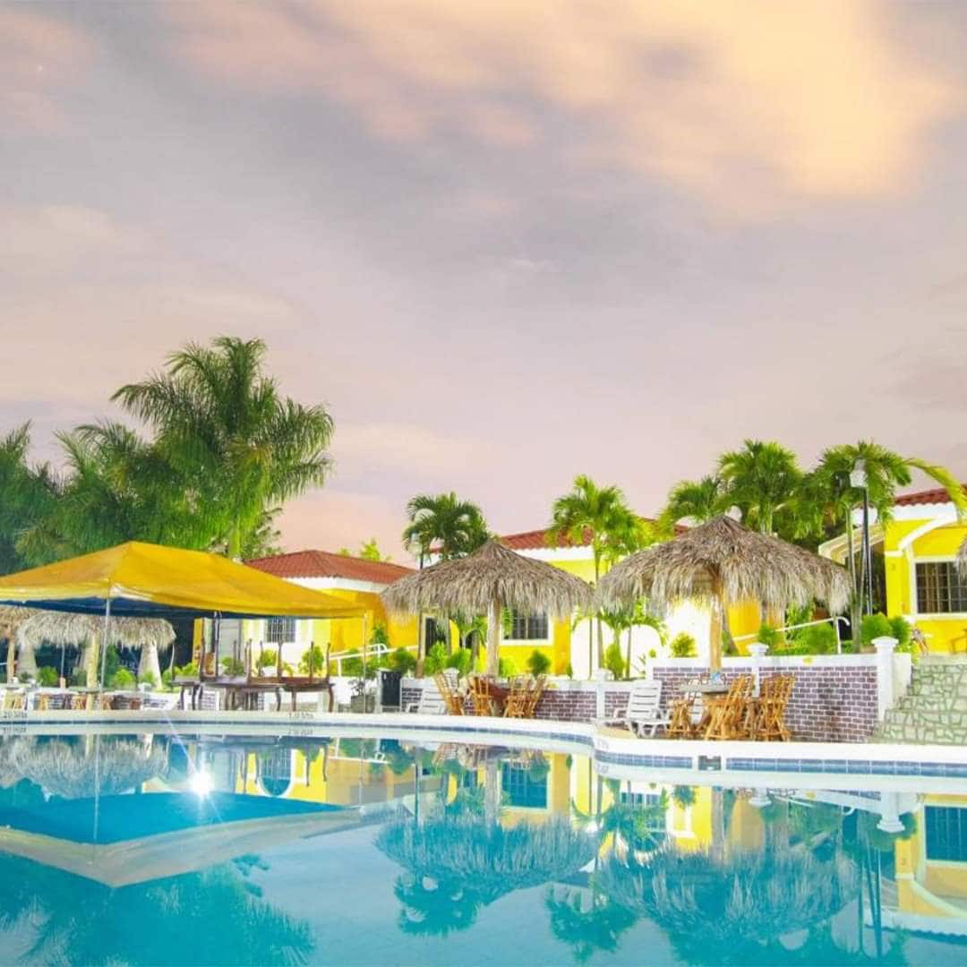 A large swimming pool reflects the evening sky, surrounded by tropical palm trees and thatched umbrellas. Colorful buildings line the background, with a covered seating area offering space for relaxation and gatherings.