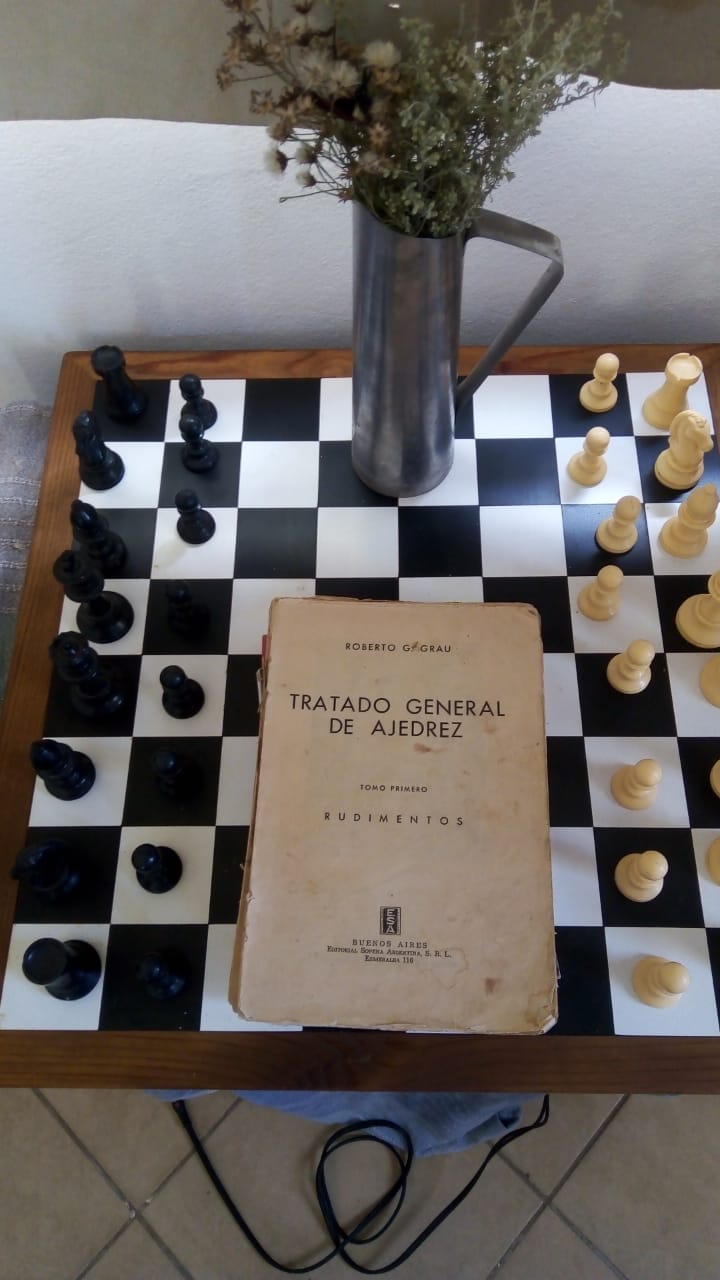 A wooden chessboard is displayed with black and white pieces positioned for a game. A chess book titled 'Tratado General de Ajedrez' rests open on the table, accompanied by a metal pitcher and dried flowers in a small vase.