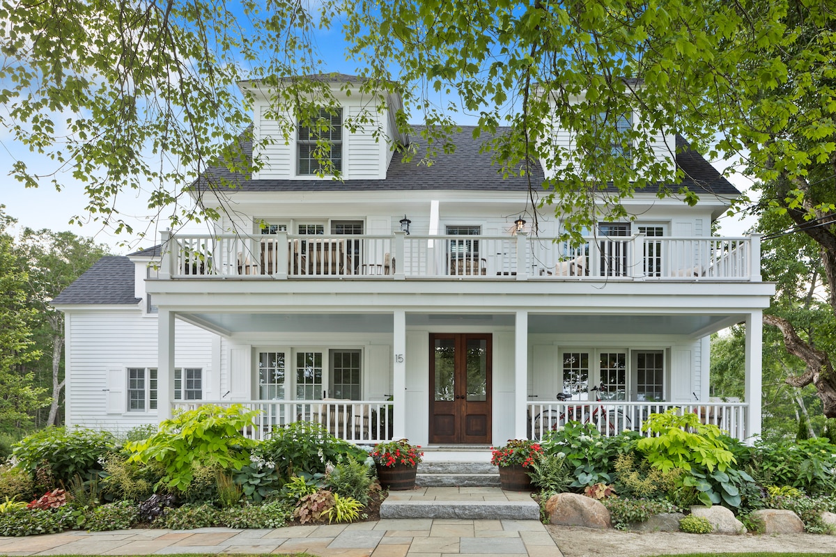 A charming two-story white house features a covered porch with railing and a central double-door entry. Lush greenery and colorful flower beds surround the property, enhancing the welcoming appearance. Large windows invite natural light, and the roof is adorned with shingles.