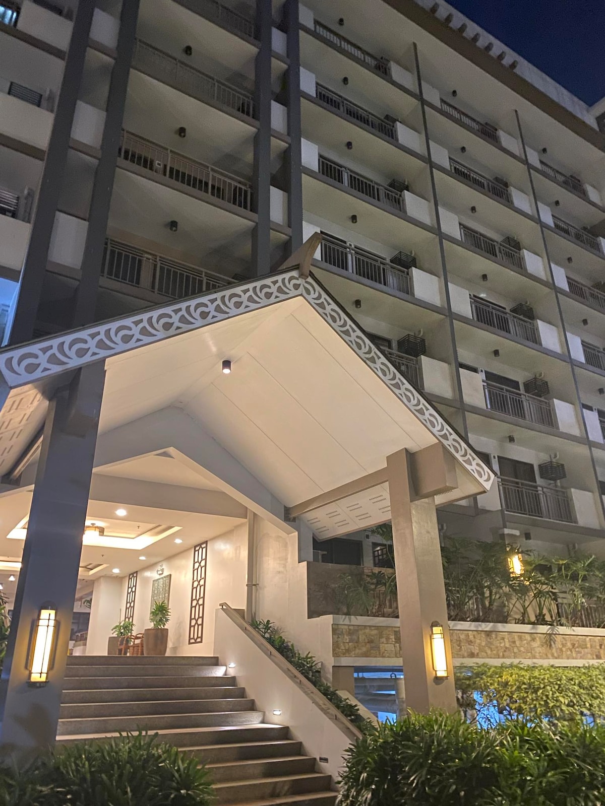 An entrance is highlighted by a covered staircase leading to a modern building. Soft lighting accentuates the architectural details, while greenery lines the path. The multi-story structure features balconies adorned with plants, creating a welcoming atmosphere.