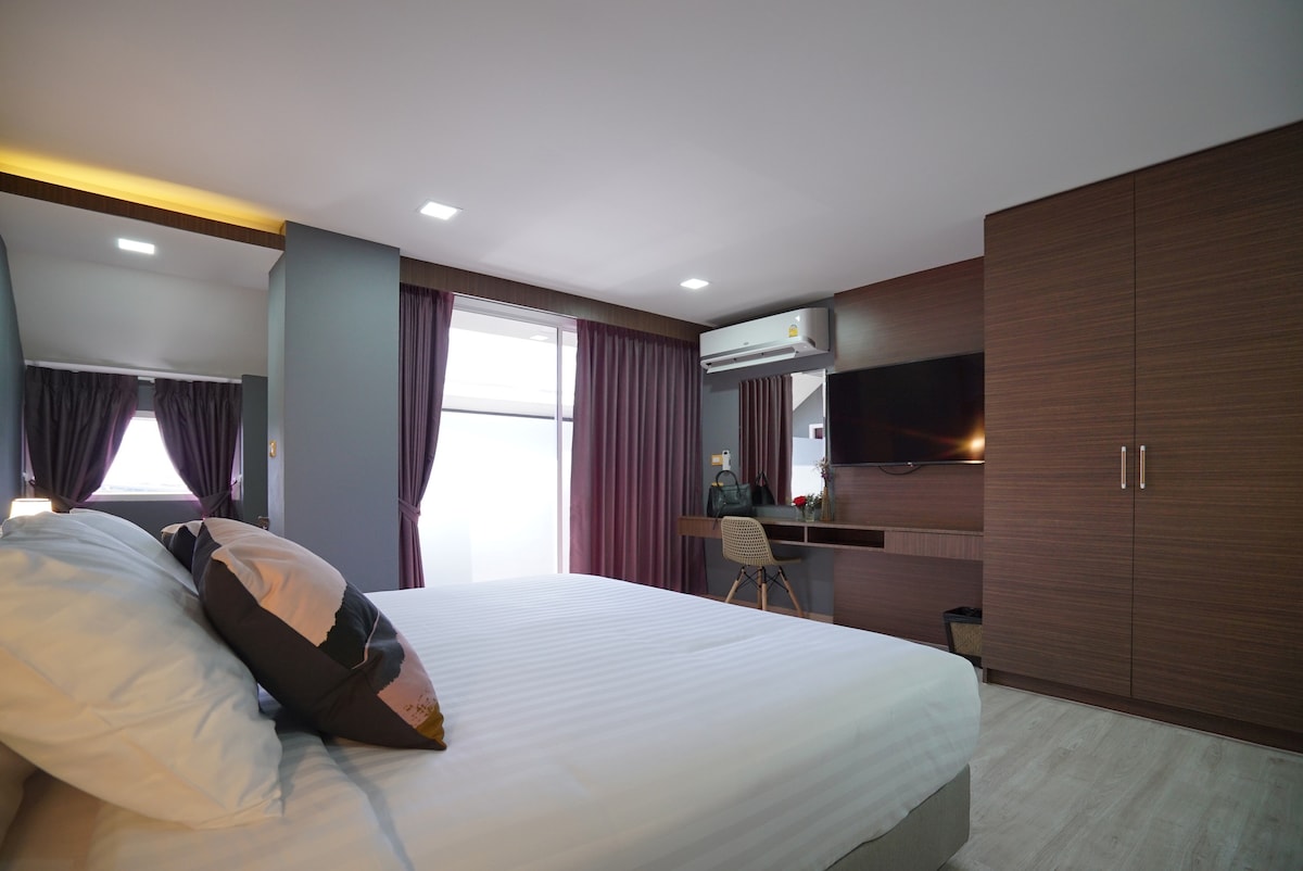 A comfortable sleeping area features a spacious bed with white linens and decorative pillows. Soft lighting illuminates the room, which includes a desk and chair adjacent to a wall-mounted smart TV. Large windows are framed by dark curtains, enhancing privacy.