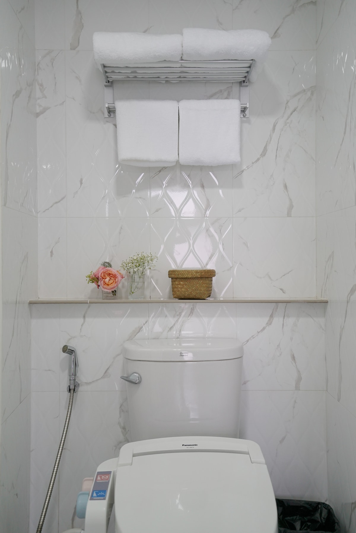 A modern bathroom area features a clean, white toilet with a sleek design. Above, neatly folded towels are stored on a rack. A small shelf displays a basket and a floral arrangement, providing a touch of color against the tiled wall.