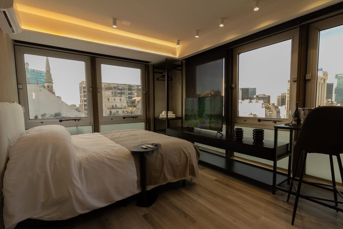 A well-lit studio is presented with large windows offering a city view. A spacious bed is featured, complemented by a simple side table. A sleek wooden console is visible, providing additional space for items. Soft tones and modern furnishings create a comfortable space.