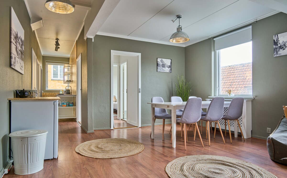 A communal dining area features a sleek white table surrounded by several light gray chairs. A refrigerator and seating area are visible in the background. Natural light streams through a window adorned with minimalistic decor, highlighting the warm wooden flooring.