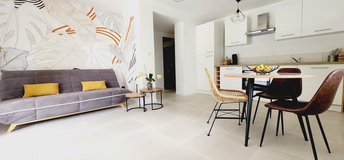 A modern living area features a gray sofa with yellow cushions, a round wooden table with four chairs, and a sliding glass door. Natural light illuminates the space, which is decorated with a wall art design. A small coffee table adds to the relaxed atmosphere.