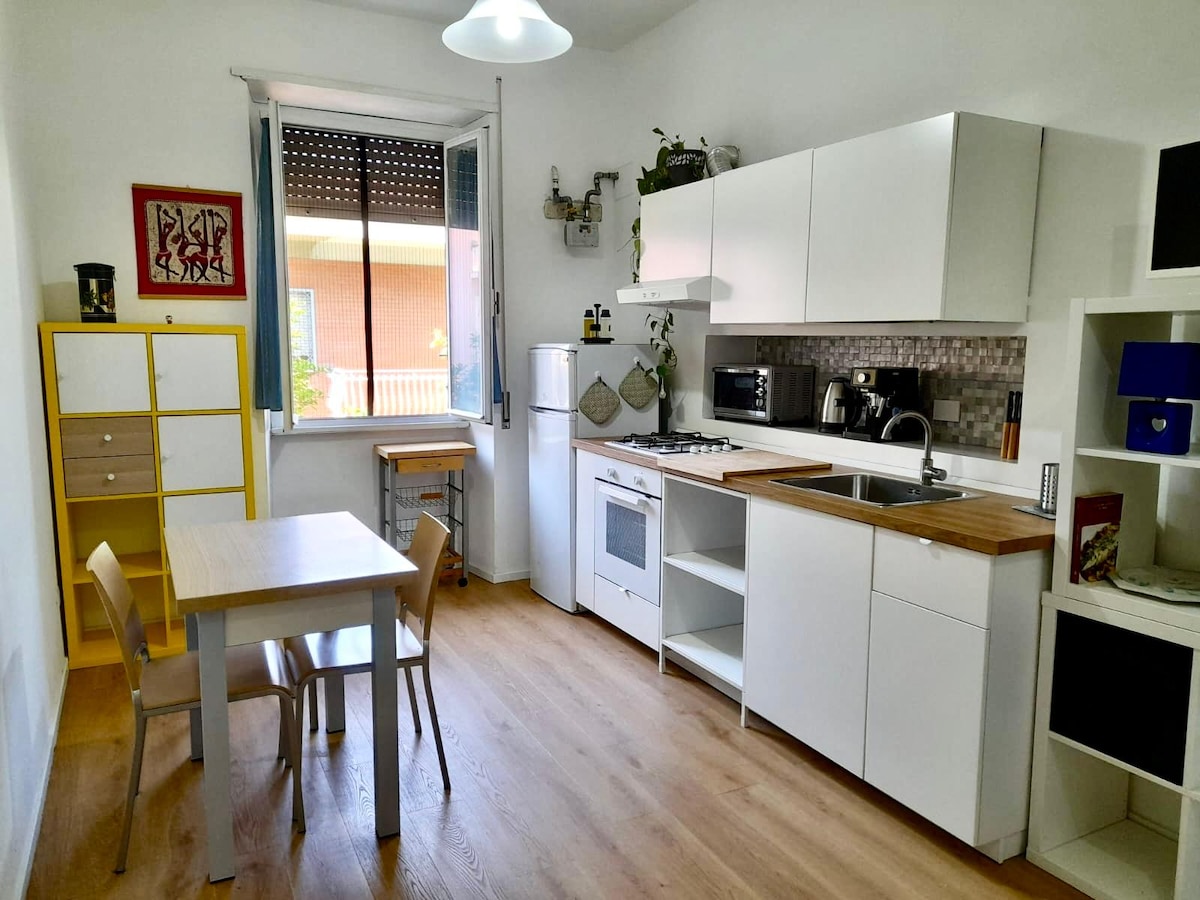 A modern kitchen space features sleek white cabinetry and a wooden countertop. A small table with four chairs is positioned near a window, allowing natural light in. Essential appliances, including a refrigerator and a stove, are neatly arranged for convenient meal preparation.