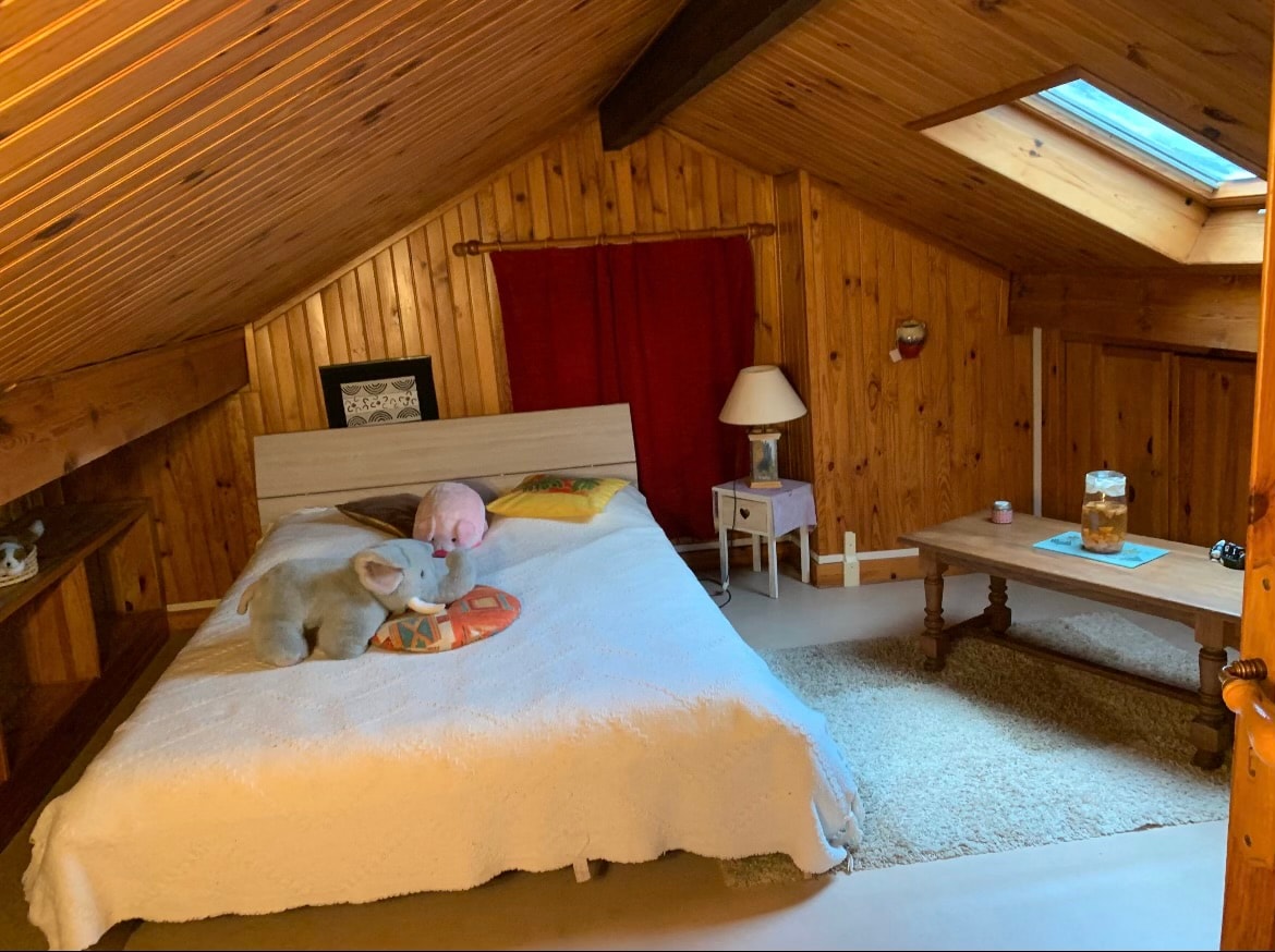 A cozy attic bedroom features wooden paneling and a slanted ceiling. A spacious bed is adorned with a light quilt and two plush toys. A small lamp sits on a side table next to the bed, while a coffee table offers refreshments. Natural light enters through a skylight.