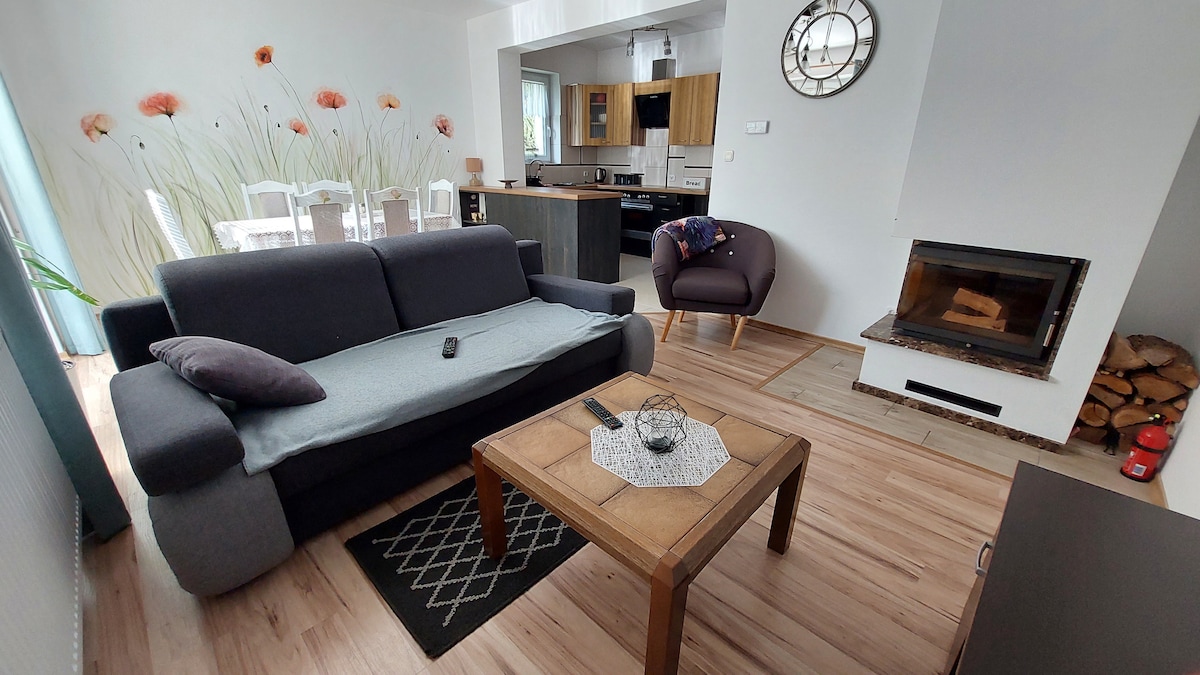 A cozy living area features a dark gray sofa with a light throw, accompanied by a brown wooden coffee table. A modern armchair sits nearby. A fireplace is positioned against the wall, while a dining table with white chairs is visible in the background, adorned with floral wall art.