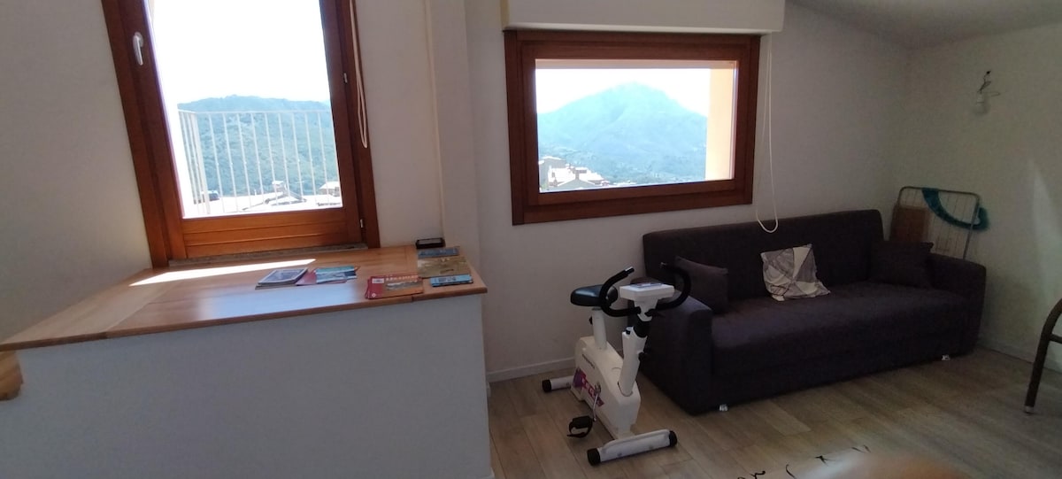 A bright and airy living space features a large window offering mountain views. A comfortable sofa is positioned beside a wooden surface with magazines. A stationary bike is also present, and light-colored flooring contributes to the room's openness.