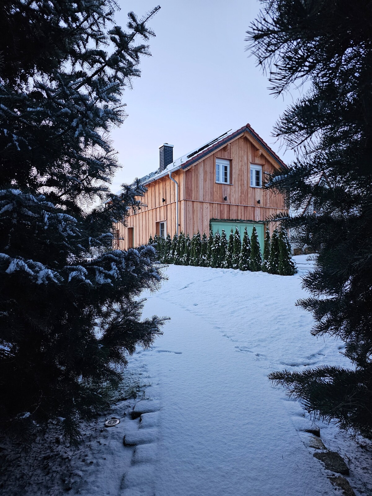 A picturesque wooden house is visible through snow-covered evergreen trees. The exterior features a warm wood finish and a light green door, framed by neatly trimmed shrubs. A pathway leads to the entrance, with fresh snow blanketing the ground.
