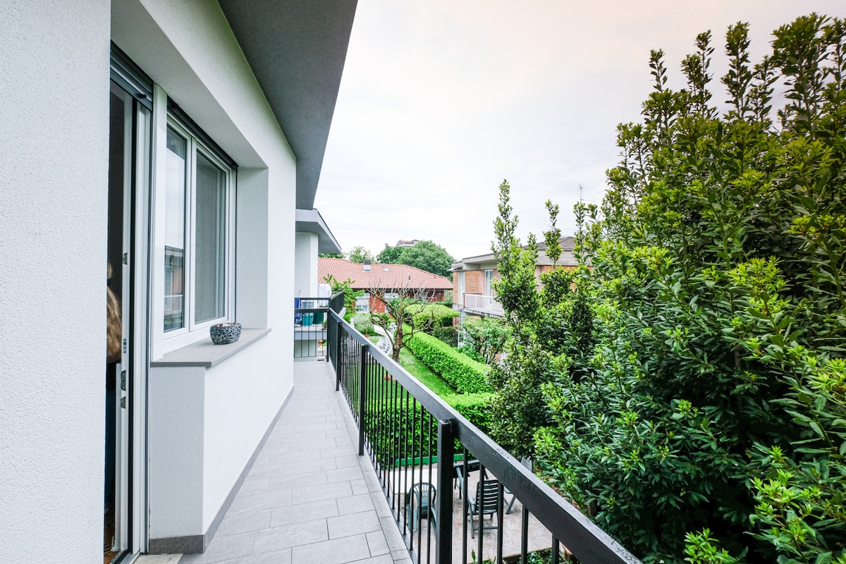 A spacious balcony extends from the living area, featuring a railing overlooking a lush green garden. Potted plants adorn the edges, and comfortable seating is visible on the patio below, creating an inviting outdoor space surrounded by greenery.