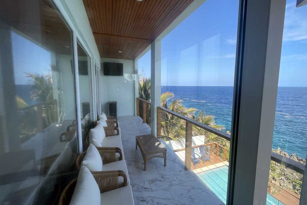 A spacious balcony offers stunning ocean views, featuring comfortable seating with white cushions and a wooden coffee table. The area is framed by large glass windows, creating an open feel. Lush greenery is visible along the coastline, enhancing the soothing atmosphere.