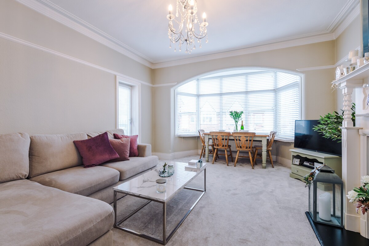 An open-plan living and dining area is bathed in natural light from large windows. A comfortable beige sofa with decorative pillows faces a glass coffee table. A wooden dining table with chairs is positioned nearby, enhancing the inviting atmosphere of the space.