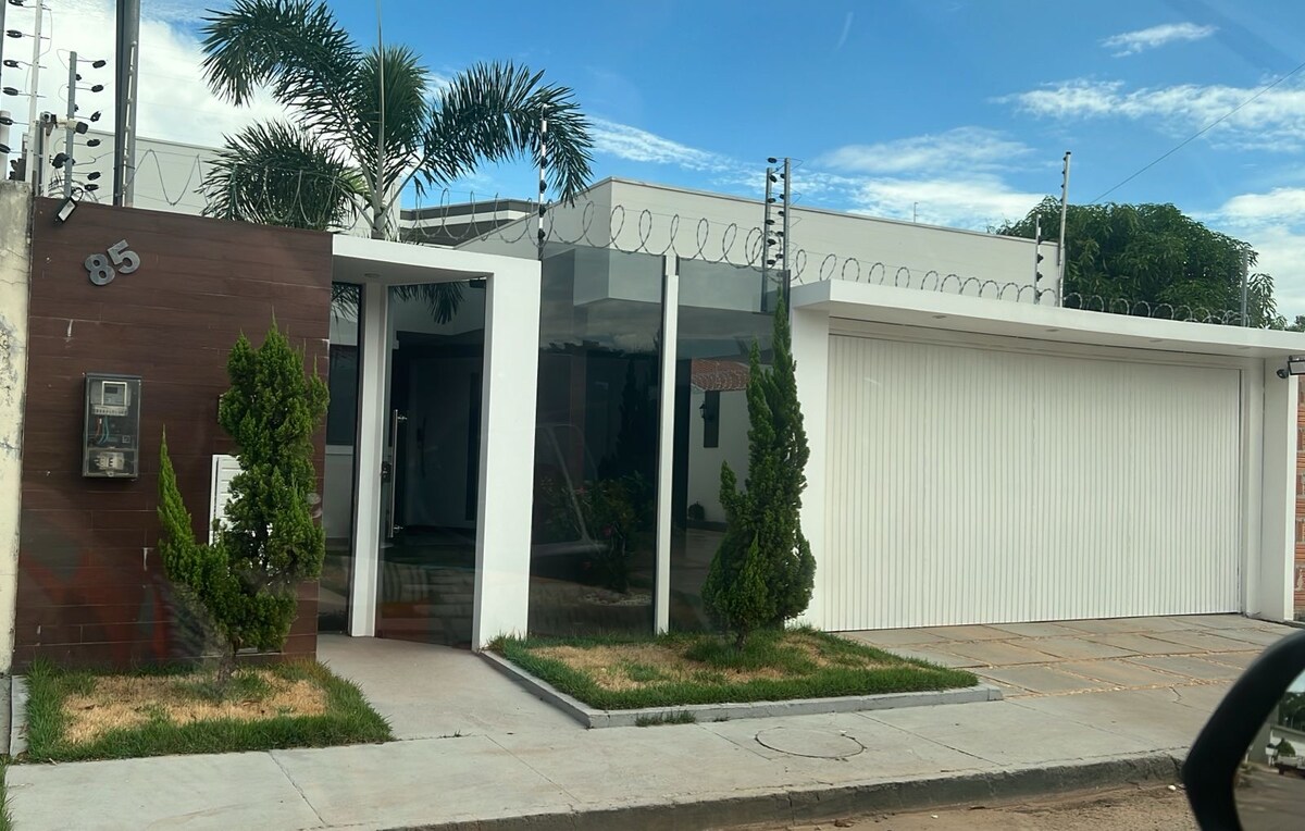 The exterior of a modern home is showcased, featuring a combination of glass and wooden elements. Lush greenery frames the entrance, while a paved pathway leads to the entrance. A garage with a white door is visible to the right side of the property.