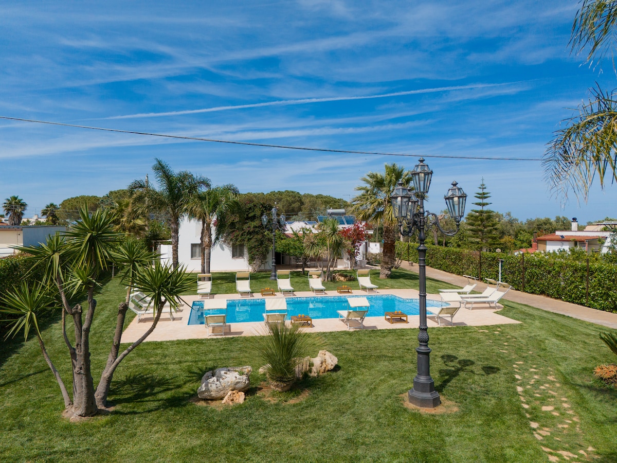 An outdoor view features a well-maintained pool surrounded by a spacious lawn. Sun loungers are arranged around the pool, and green palm trees provide shade. A decorative lamp post stands in the foreground, adding charm to the serene landscape.