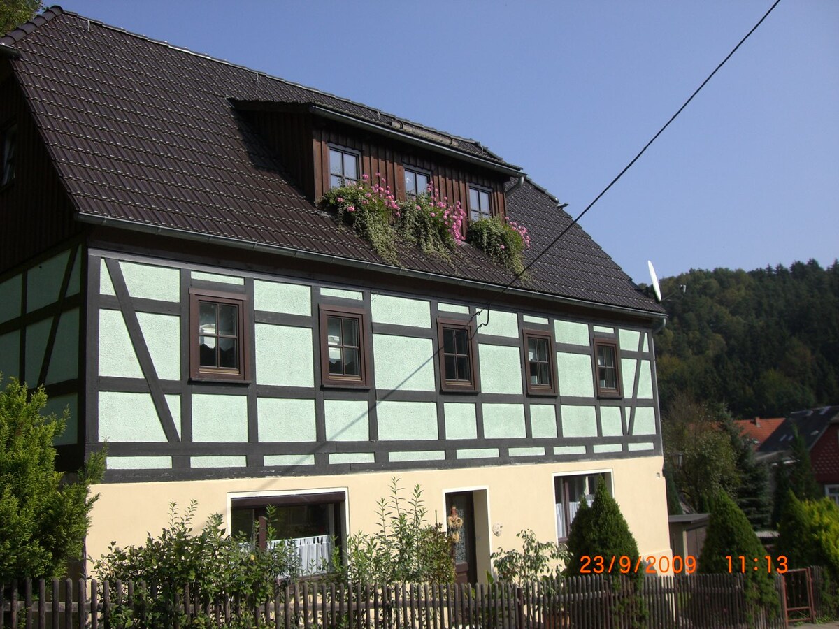 A historic half-timbered building features a green and cream facade with decorative plants adorning the upper windows. The structure includes multiple windows and a pitched roof, set against a backdrop of greenery. A small front garden with shrubs is visible, enhancing the natural surroundings.