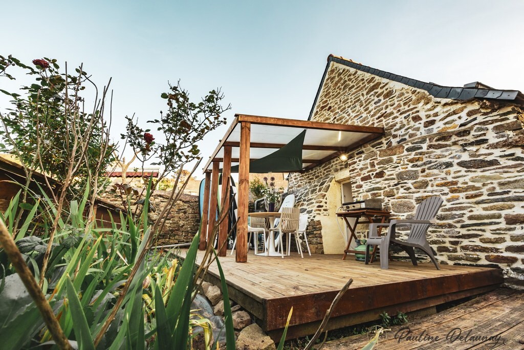 A wooden terrace is presented, featuring a sheltered area with seating and a dining table. Surrounding greenery and textured stone walls complement the outdoor space, enhancing the inviting ambiance for relaxation and meals outdoors.