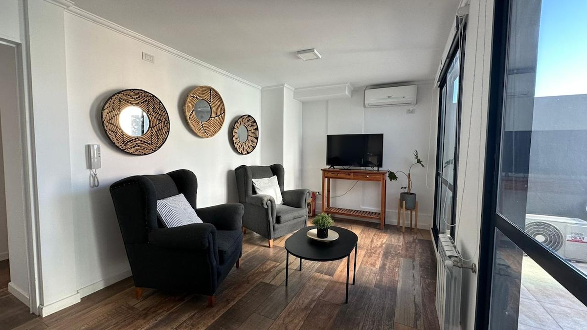 A living area features two comfortable chairs facing a modern flat-screen TV. A small round table is placed in the center, while decorative mirrors adorn the walls. Natural light is streamed in through large windows, enhancing the welcoming atmosphere.