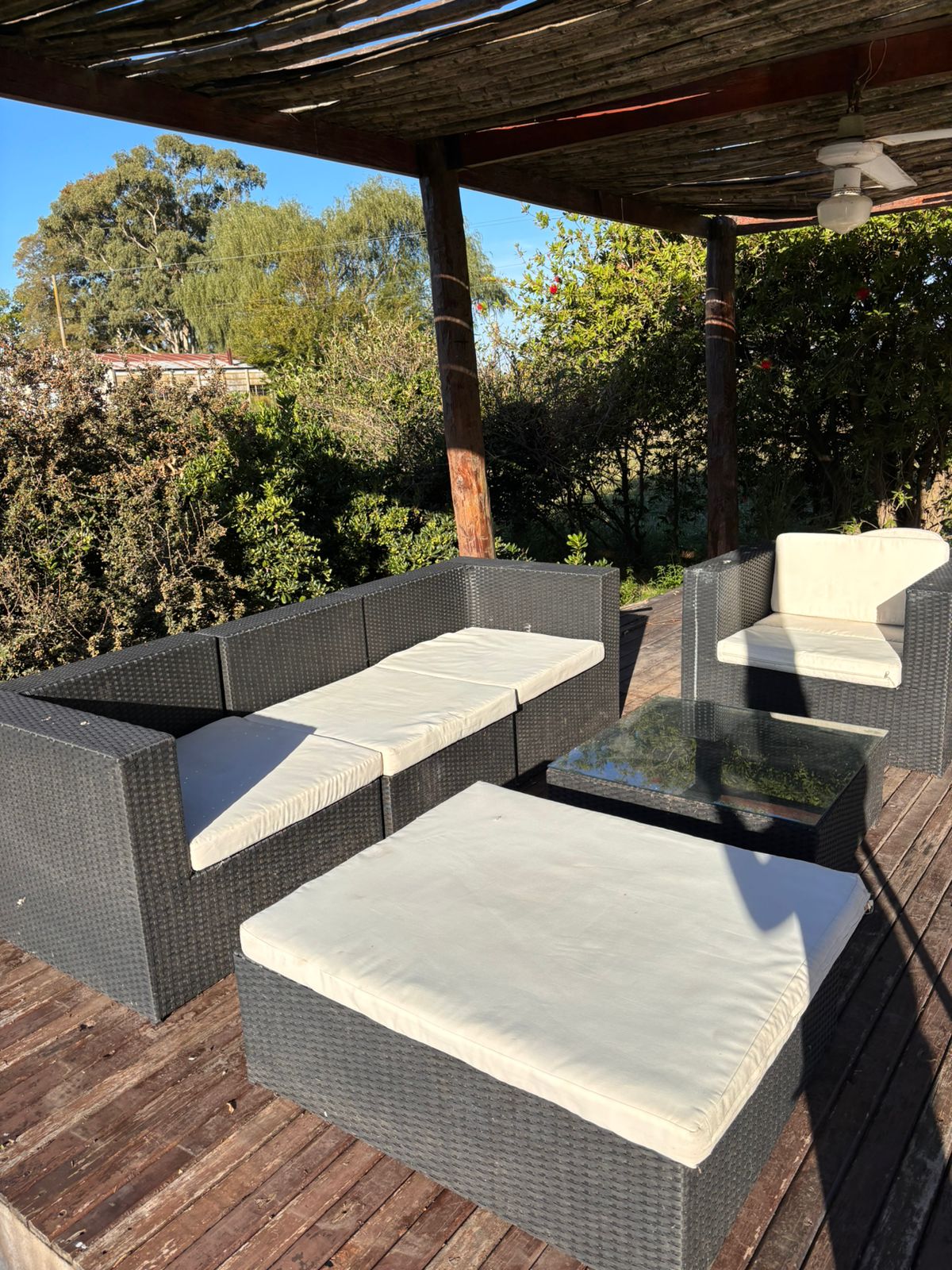 An outdoor seating area is featured, with a dark wicker furniture set including a sectional sofa and a low table. Cushions in neutral tones are placed on the seating. The space is partially shaded under a wooden pergola, surrounded by greenery.