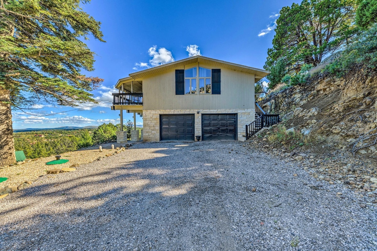The exterior of the house is shown, featuring a two-story structure with a light-colored facade. A spacious driveway leads up to a double garage with dark doors. Surrounding trees and a rocky terrain contribute to the natural setting, with a clear blue sky overhead.