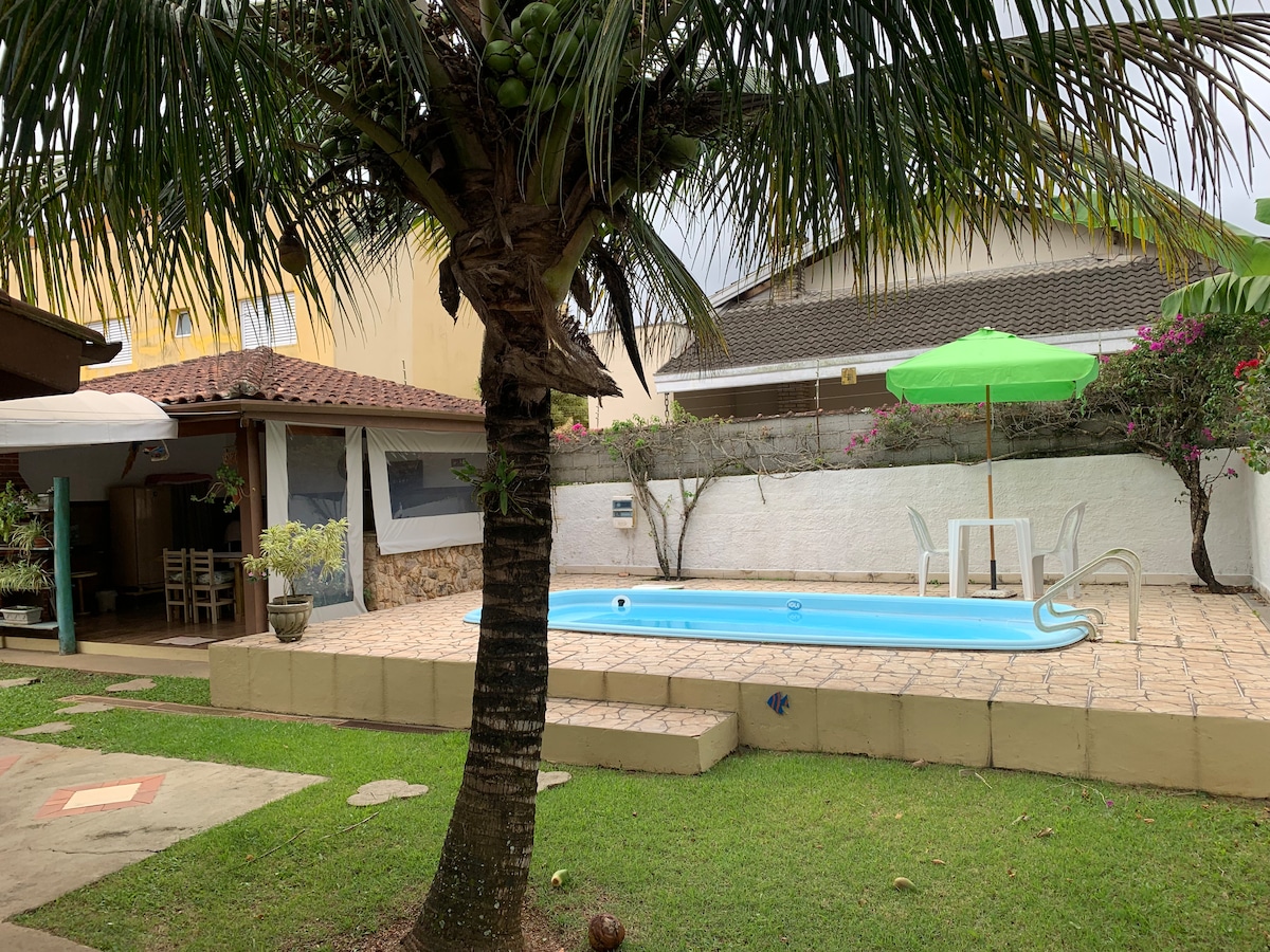 A private pool is surrounded by a patio area, featuring a green umbrella above a small table with chairs. Lush grass and a palm tree provide a natural setting, while a covered outdoor space is visible in the background.