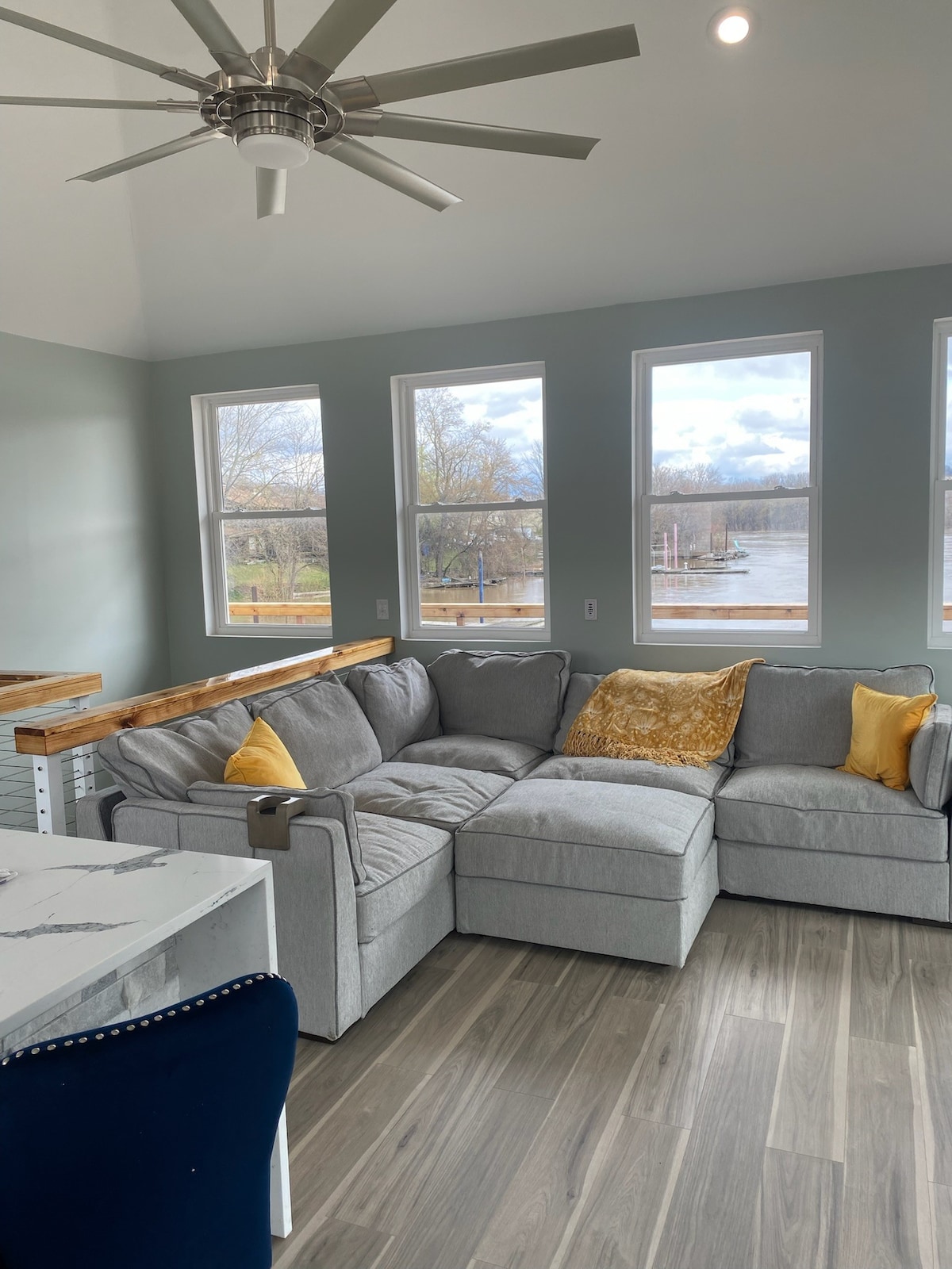 An open living area is highlighted by a spacious gray sectional sofa adorned with vibrant yellow pillows. Large windows allow natural light to illuminate the space while offering views of the river outside. A simple dining table is visible in the foreground.